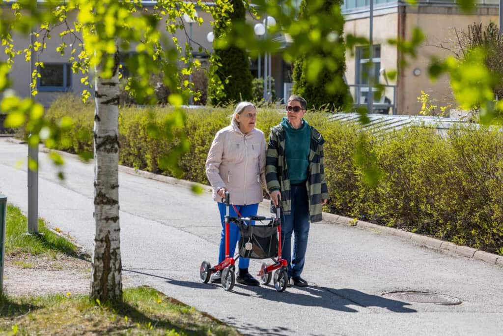 Iäkäs nainen ja mies kävelevät ulkona, nainen käyttää rollaattoria ja mies tukena, ympärillä vihreää.